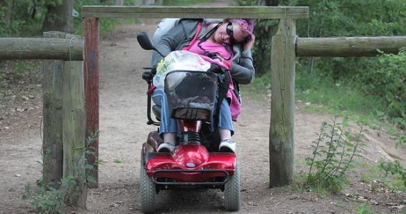 Rollstuhlfahrerin auf einem Waldweg bückt sich unter einem Hinderniss