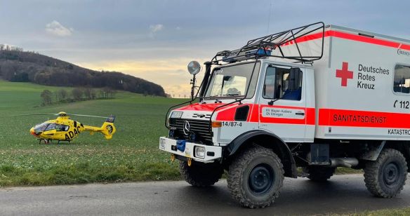 Ein geländegängiges Fahrzeug des DRK steht vor einer Wiese, im Hintergrund ist ein ADAC Hubschrauber zu sehen