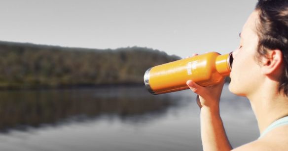 Mann steht in der Natur an einem See und trinkt Wasser aus einer Flasche