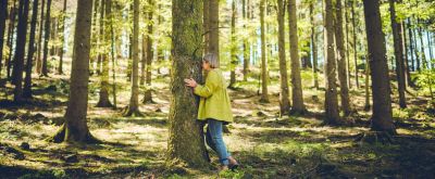 Frau steht im Wald, sie berührt mit den Händen und ihrem Gesicht einen Baumstamm
