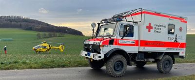 Ein geländegängiges Fahrzeug des DRK steht vor einer Wiese, im Hintergrund ist ein ADAC Hubschrauber zu sehen